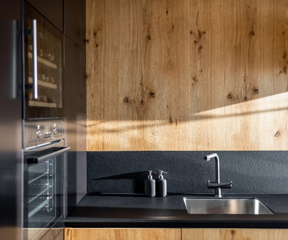 Cuisine moderne : évier inox, robinet noir, plan de travail sombre et mur en bois clair sous la lumière naturelle.