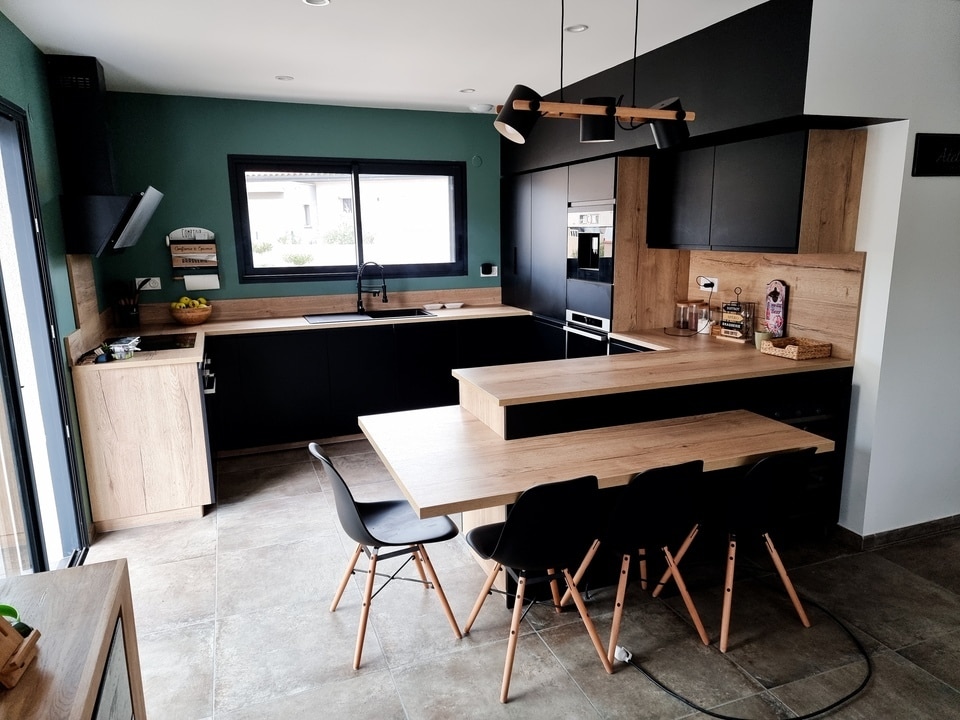 Cuisine design noir et bois avec mur vert sombre, îlot central prolongé en table et chaises style scandinave.