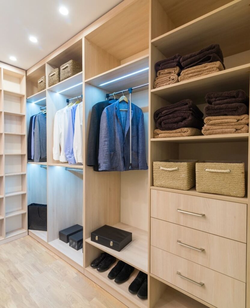 Dressing moderne en bois clair avec vêtements suspendus, étagères de serviettes pliées, tiroirs et éclairage LED intégré.
