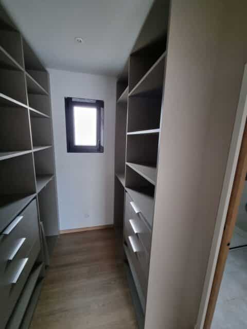Dressing sur mesure gris avec étagères, tiroirs et parquet clair, éclairé par une petite fenêtre au fond.